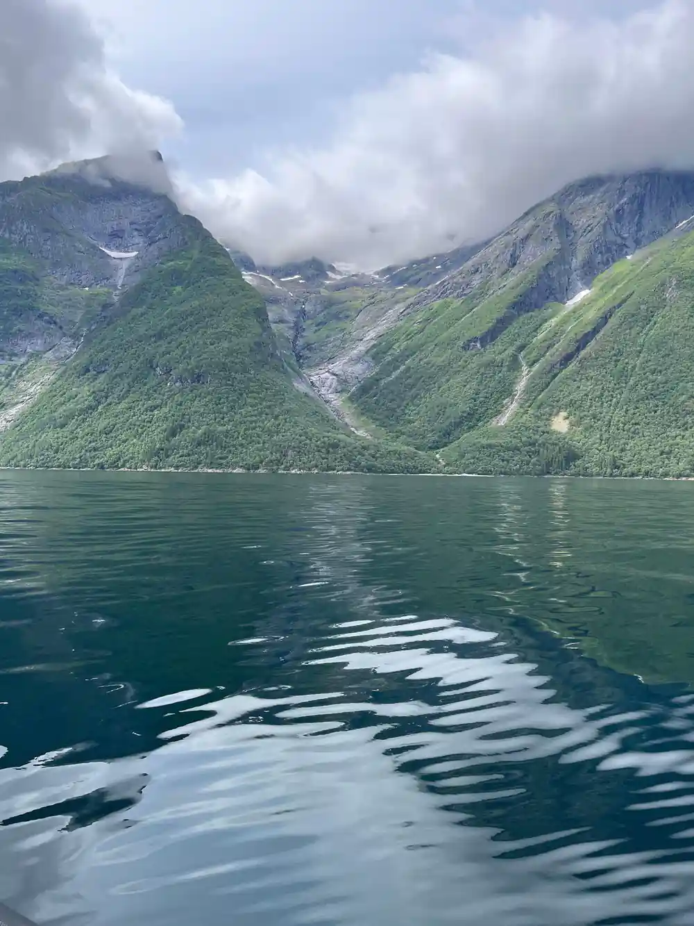 A green mountain wall with snow above a still fjord