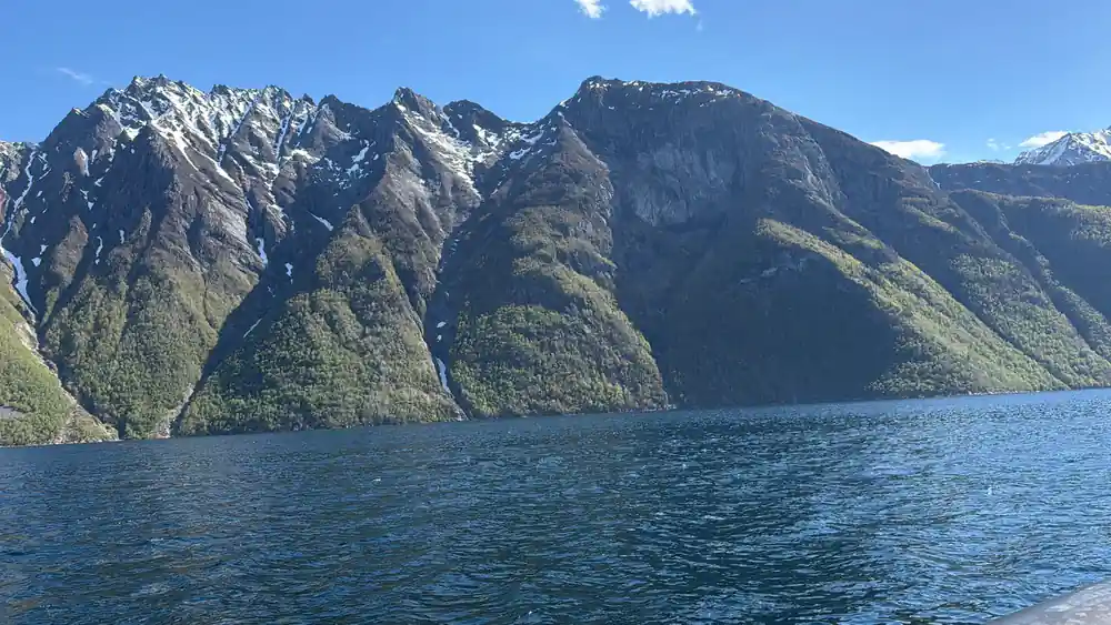 A steep dark peak with snow above deep blue water